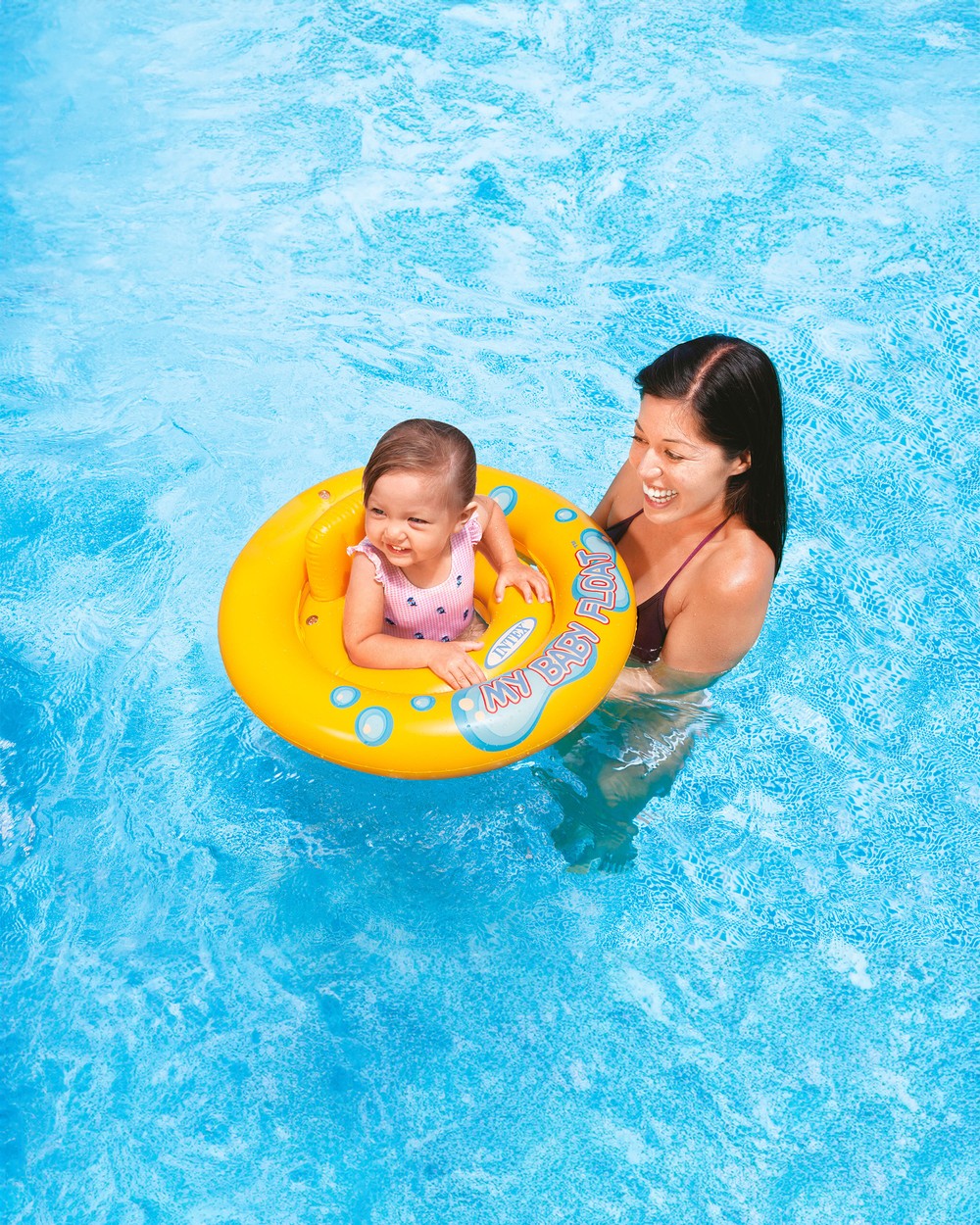 A happy baby sitting in an Intex My Baby Float ring, held by mum in a bright blue pool